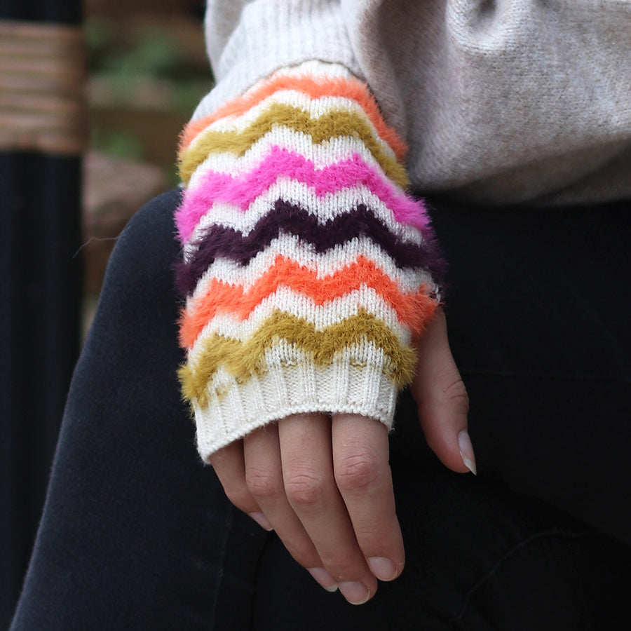 A hand wearing a knitted fingerless glove with chevron stripes in pink, purple, orange, mustard and white rests on a black surface—ideal for fans of colourful winter accessories.