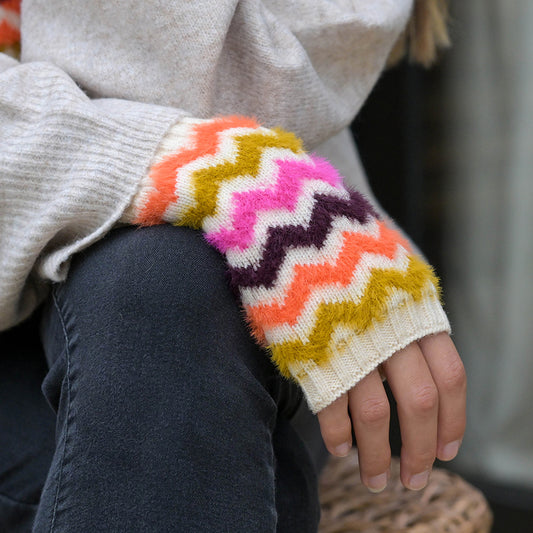 A person wearing a beige jumper with colourful zigzag-patterned cuffs and dark trousers rests their hand, covered in super soft Chevron mittens, on their knee.