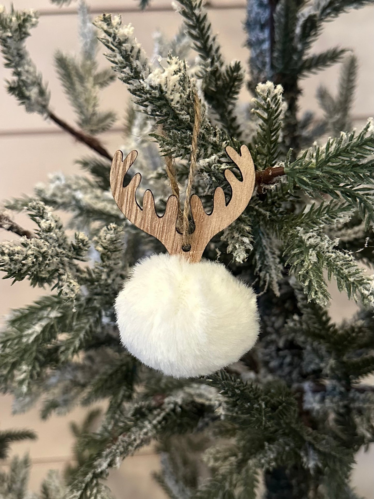 A Deer Pom Pom Christmas tree bauble, shaped like a white fluffy ball with wooden antlers, hangs on a snow-dusted branch—ideal for festive holiday décor.