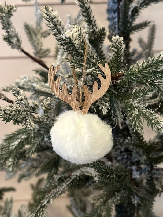 A Deer Pom Pom Christmas tree bauble, shaped like a white fluffy ball with wooden antlers, hangs on a snow-dusted branch—ideal for festive holiday décor.