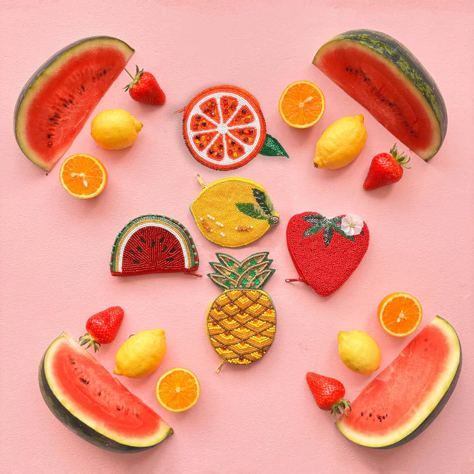 Slices of watermelon, lemons, strawberries, and oranges encircle decorative fruit-shaped coin purses—including a pineapple-shaped purse—on a pink background, creating a quirky design that's an ideal vegan-friendly gift.