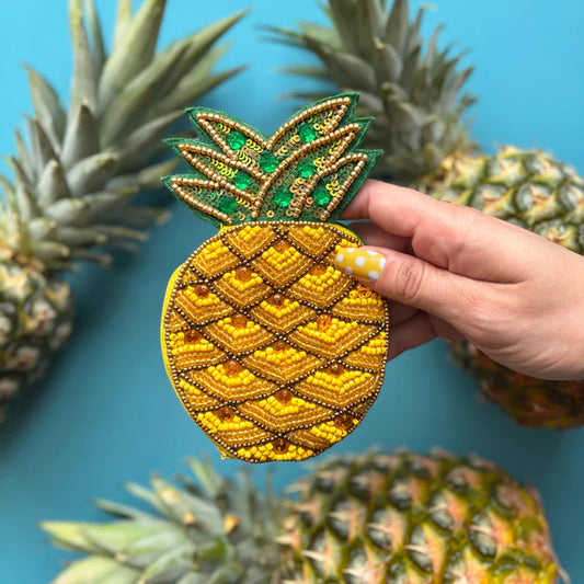 A hand with yellow polka-dot nail art holds a quirky, pineapple-shaped purse, surrounded by real pineapples on a blue background—a fun, vegan-friendly present idea.