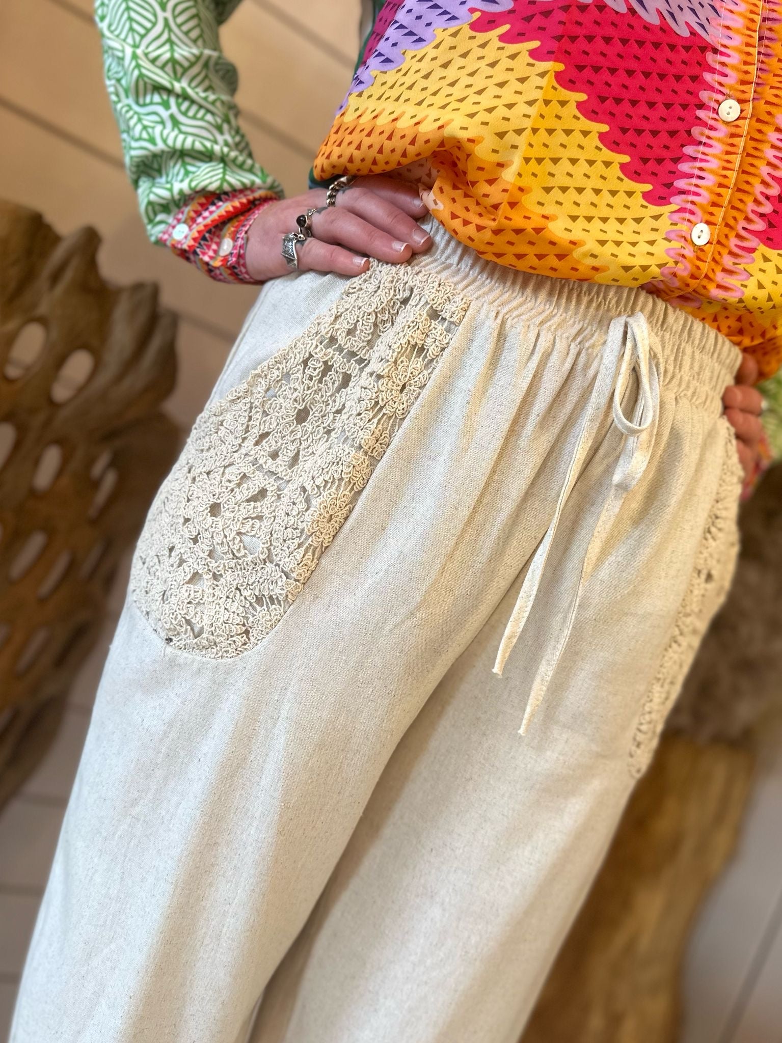 A person wearing light-coloured linen trousers with crocheted pockets and a funky statement shirt stands indoors, perfectly dressed for festival season.