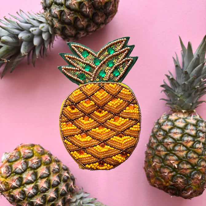 A beaded pineapple brooch with a quirky design sits on a pink background, bordered by four real pineapples at the edges—a perfect vegan-friendly gift for fruit enthusiasts.