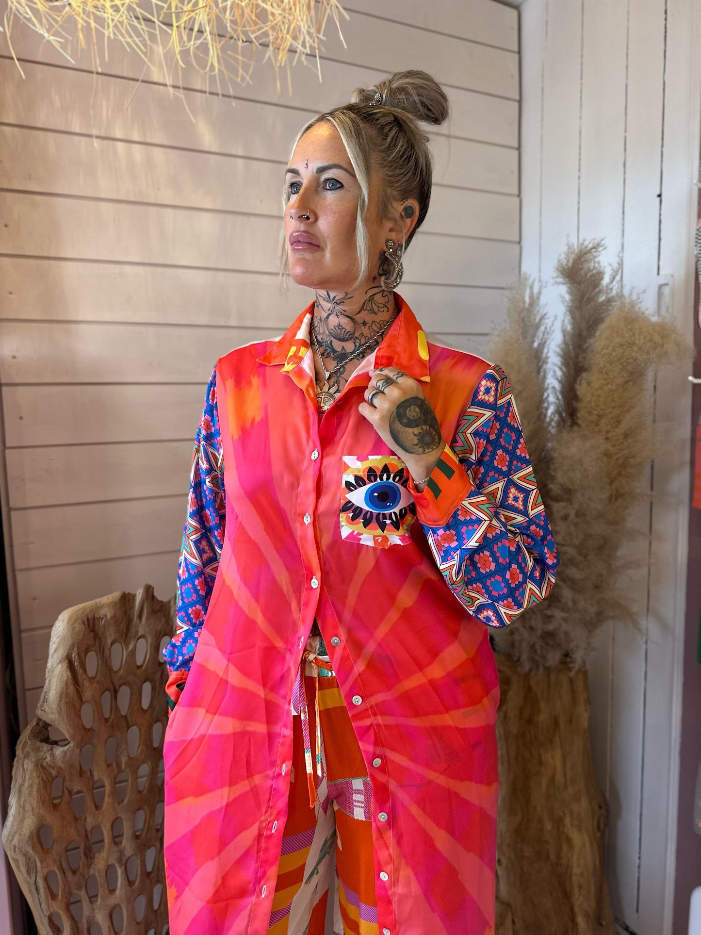 A woman with tattoos and facial piercings stands indoors, wearing a brightly patterned Eye Kaftan with a unique design. She gazes to the side, with neutral-coloured décor in the background.