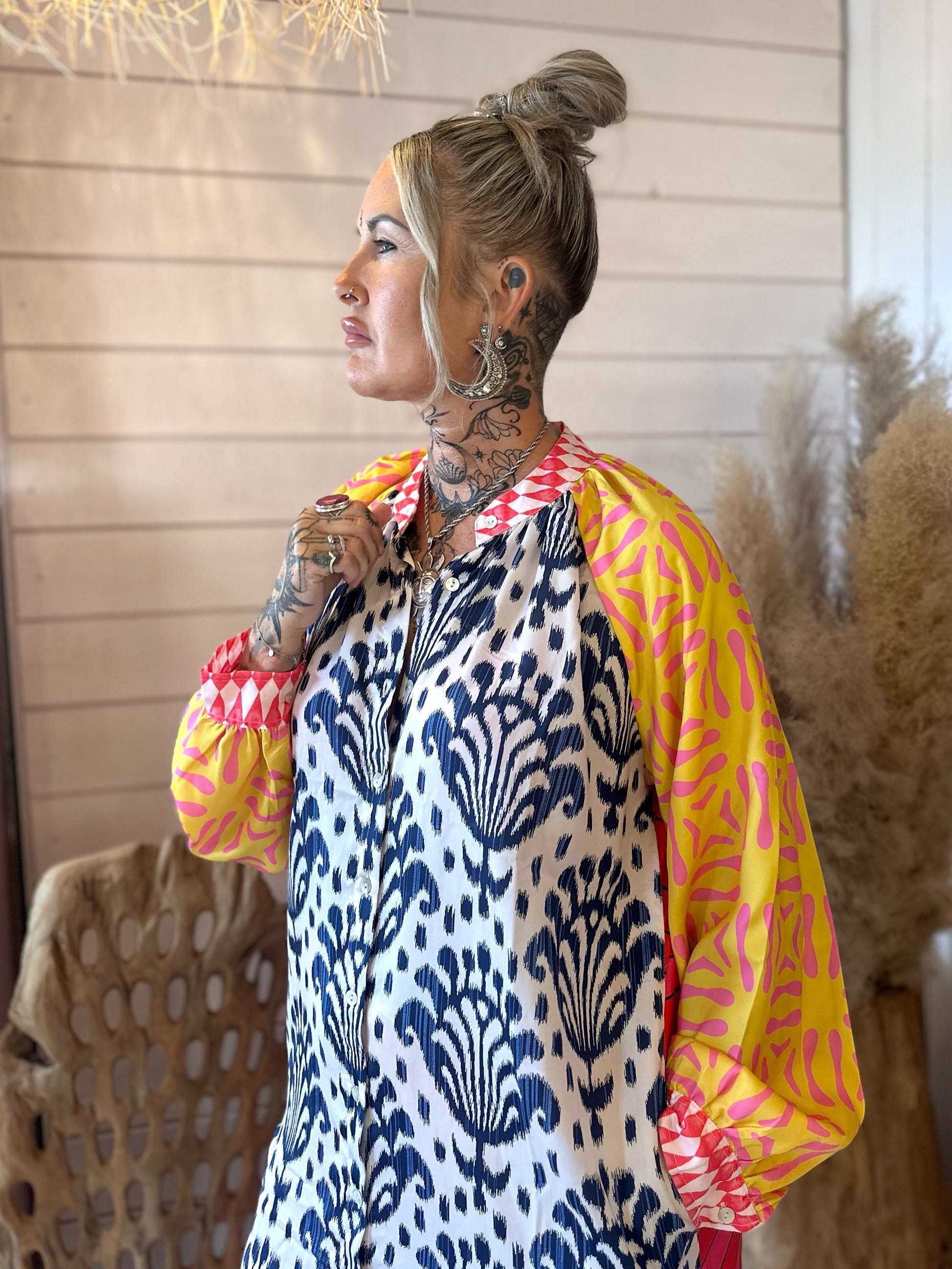 A woman with tattoos and blonde hair in a bun is wearing a patterned dress with yellow and pink sleeves, standing indoors near beige pampas grass.