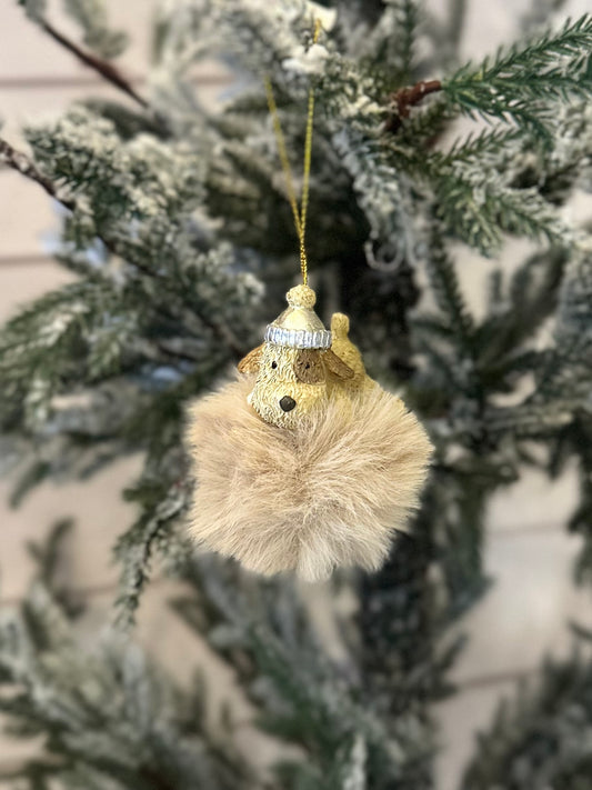 A dog-themed Christmas tree decoration shaped like a fluffy brown sphere with a small dog's head wearing a winter hat, ideal festive décor for dog lovers, hanging from a snow-covered tree branch.