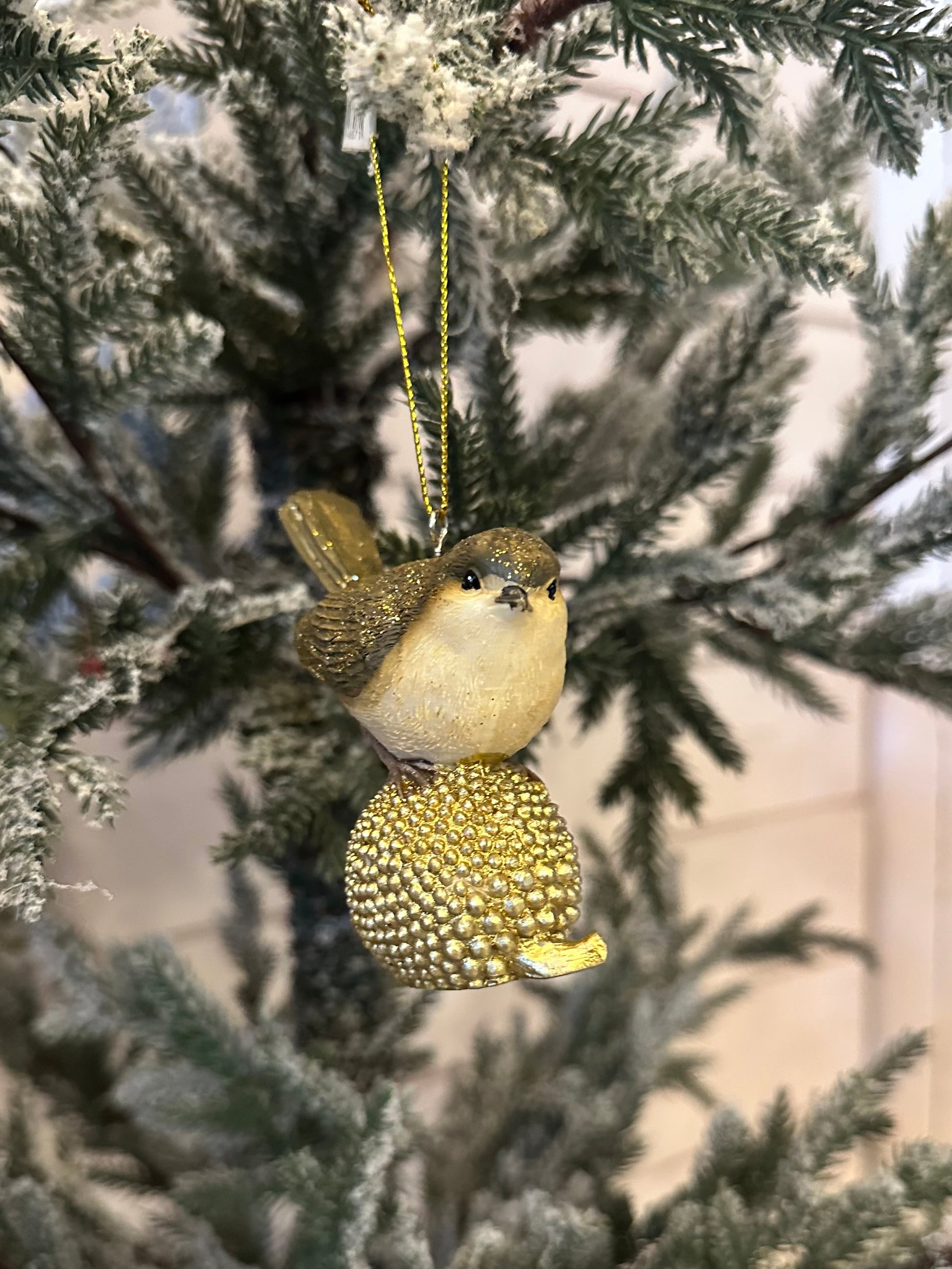 A gold-dusted bird ornament with a golden textured sphere hangs from a snow-dusted artificial Christmas tree branch, bringing festive charm to your holiday décor.