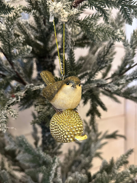 A gold-dusted bird ornament with a golden textured sphere hangs from a snow-dusted artificial Christmas tree branch, bringing festive charm to your holiday décor.