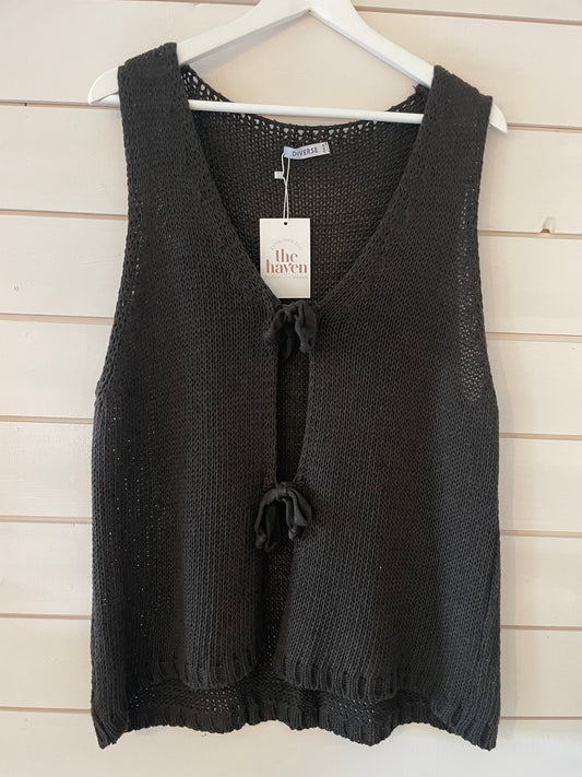 Sleeveless black knitted vegan waistcoat with two front ties, shown on a white hanger against a light wooden wall. A tag reading "the haven" is attached to the waistcoat. Ideal for versatile layering.