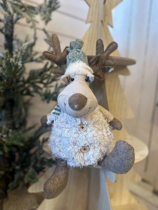 A plush Sitting Deer Christmas Decoration, featuring button details and a green hat, sits on a wooden chair before a tree-shaped ornament, making the ideal festive companion for your holiday décor.