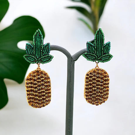 Add a touch of tropical fun to your look with these Beaded Crystal Pineapple Earrings, featuring green leaves and golden beaded fruit, beautifully displayed on a stand with luxuriant foliage in the background.