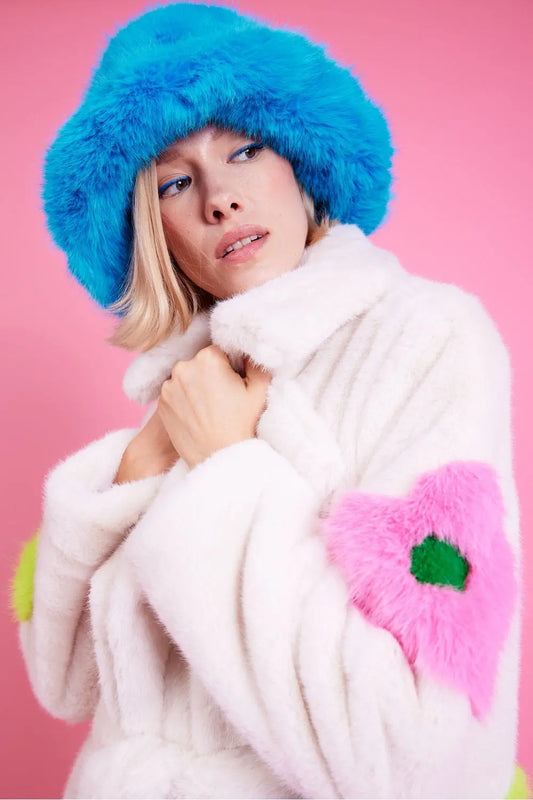 A person wearing a Blue Electric Eco Bamboo Bucket Hat with a round brim and a white faux fur coat featuring pink and green patches stands against a pink background.