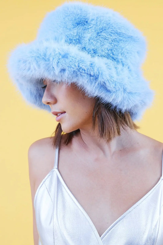 A person wearing a light blue faux fur bamboo bucket hat and a silver spaghetti strap top stands against a yellow background.