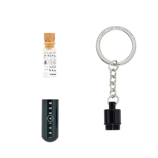 A small glass vial with a cork, a black capsule labelled BEEVIVE Bee Revival Kit, and a metal keyring with a screw-on attachment are displayed on a white background.