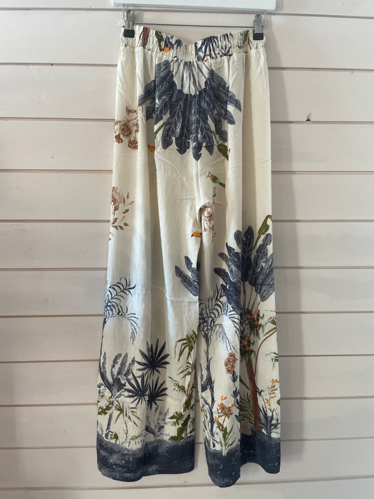 Blue jungle trousers in lightweight fabric with an elasticated waistband, showcasing a botanical print in navy, green, and brown on a cream base, hung on a clear hanger against a white wooden wall.