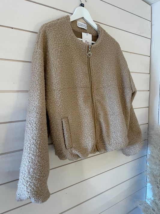 Cream bomber jacket with a relaxed fit, made from textured fleece with a zip front and long sleeves, hanging on a white hanger against a white wooden wall.