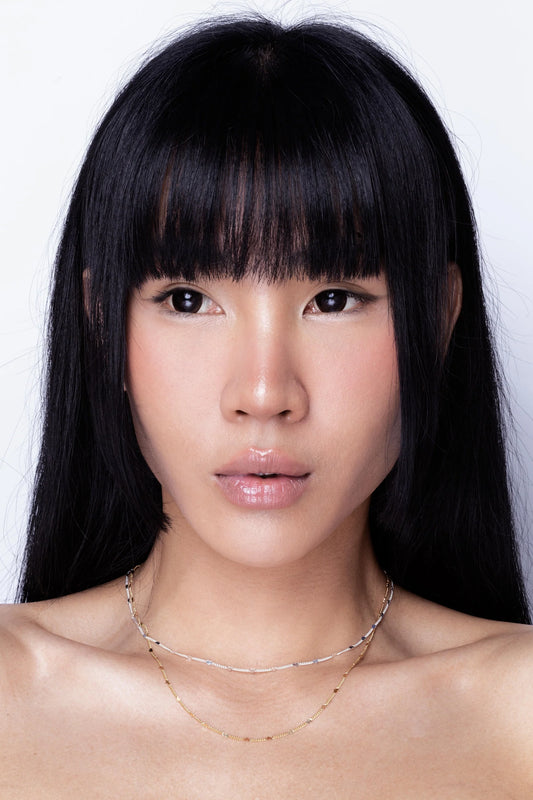 A woman with straight black hair and a fringe wears a delicate hypoallergenic necklace and a gold chain against a plain white background.