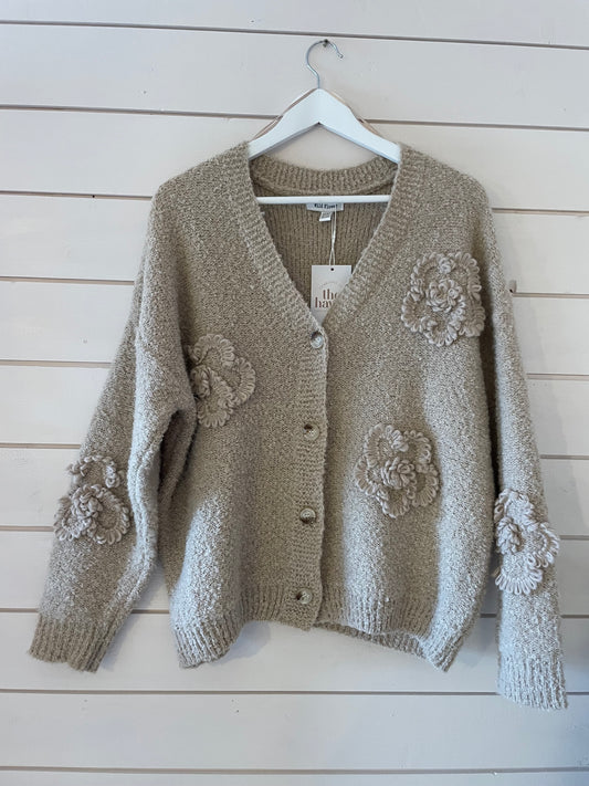 The Soft Flower Cardigan is a beige knit with large flower appliqués on the front and sleeves, made from ultra-soft fabric and shown on a white hanger against a shiplap wall.