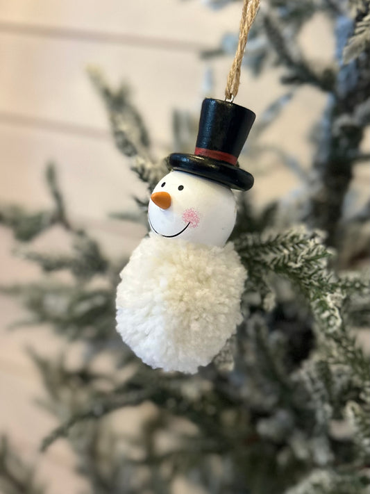 A snowman ornament wearing a black top hat hangs from a Christmas tree branch, surrounded by green needles and a light sprinkling of snow in the background.