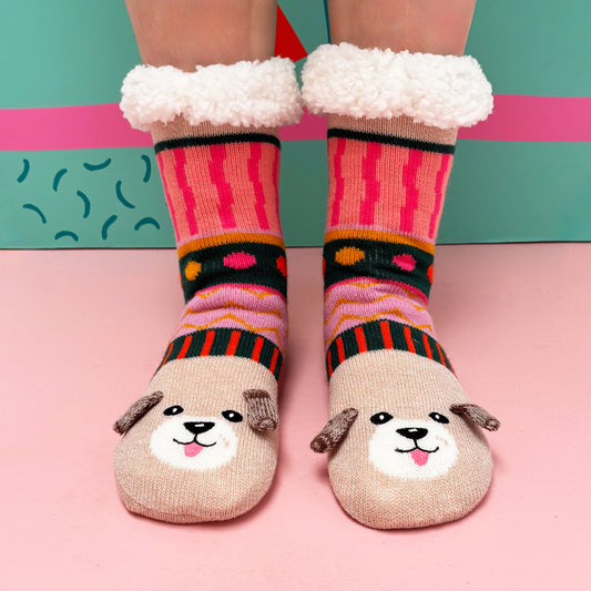 A person is wearing cosy, colourful Dog House Slipper Socks with a dog face design and fluffy cuffs, standing on a pink surface before a teal background—ideal as a gift for dog lovers.