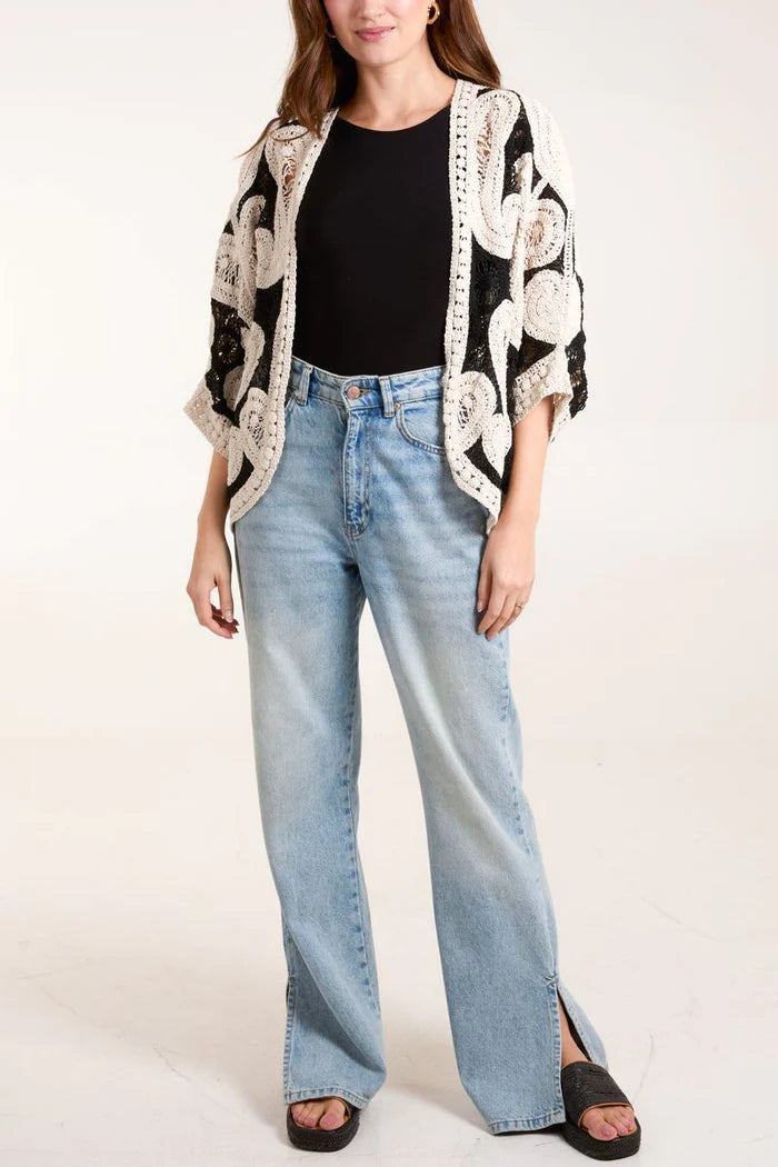 A person wearing a black top, 100% cotton paisley crochet kimono with white patterns, light blue wide-leg jeans, and black platform sandals.