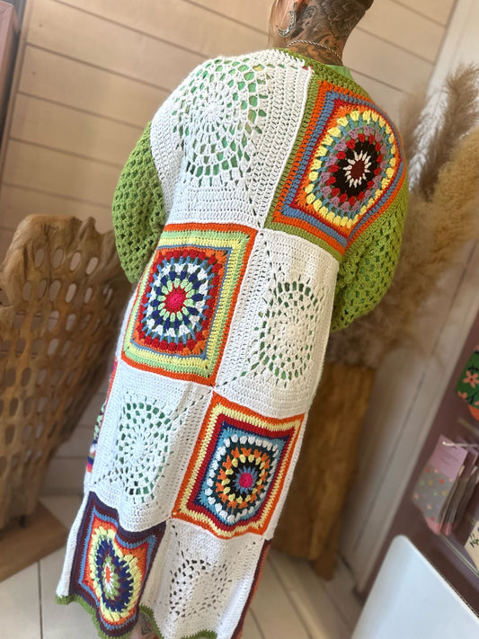 Person wearing a hand-crafted crochet long cardigan with multicoloured patchwork detailing and large square and circular patterns on the back, standing indoors.