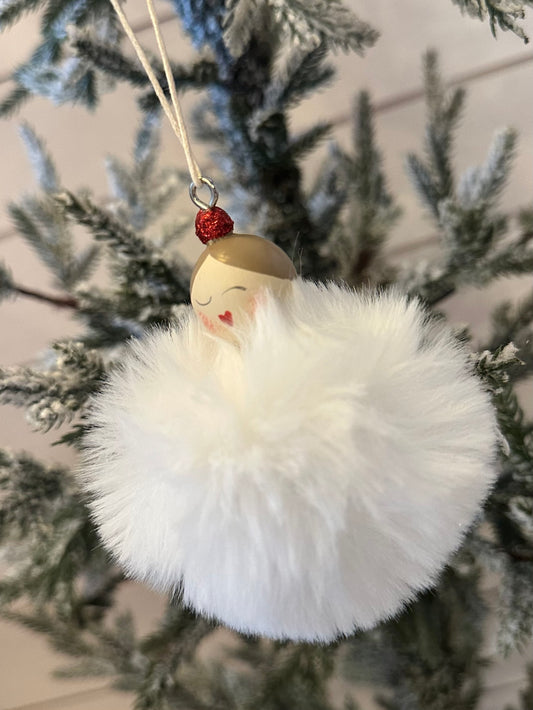 A Christmas decoration in the shape of a pom-pom style lady angel, with a painted face, red hat, and fluffy white body, hangs from a branch of a frosted tree.