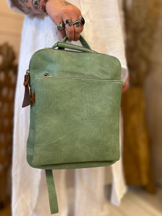 A person wearing rings and dressed in white is holding a small green rucksack with a front pocket and a top handle.