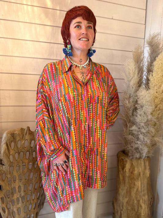 A person standing indoors wearing a vegan-friendly, multicoloured patterned Loxy Long Top and white trousers, accessorised with jewellery and blue earrings, with dried plants and wooden décor in the background.