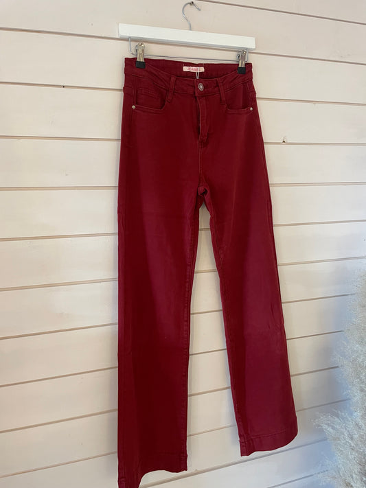 A pair of high-waisted, straight-leg wide leg red jeans hangs on a white hanger against a light wooden wall—a stylish denim piece for any wardrobe.