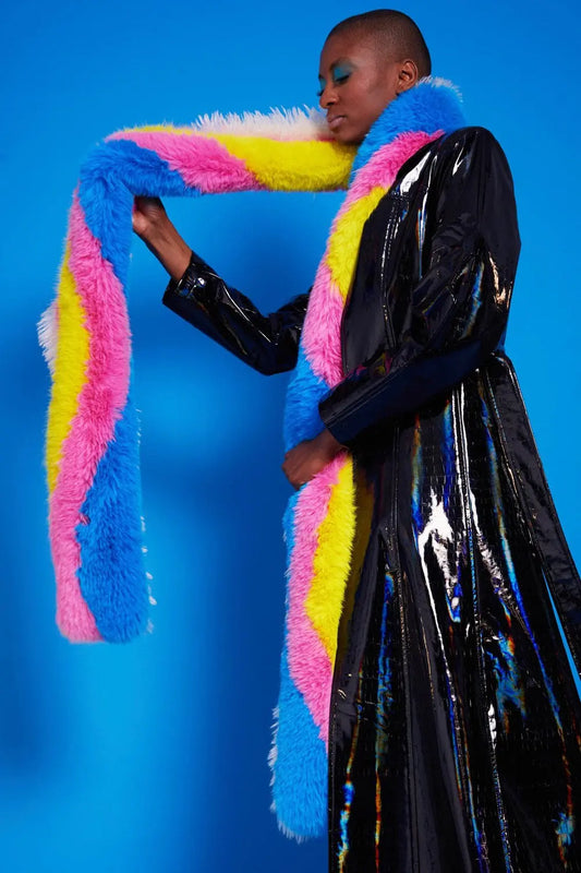 Person wearing a shiny black trench coat and a dove grey faux fur snood with blue, pink, yellow and white stripes—a chic winter accessory—posing against a blue background.