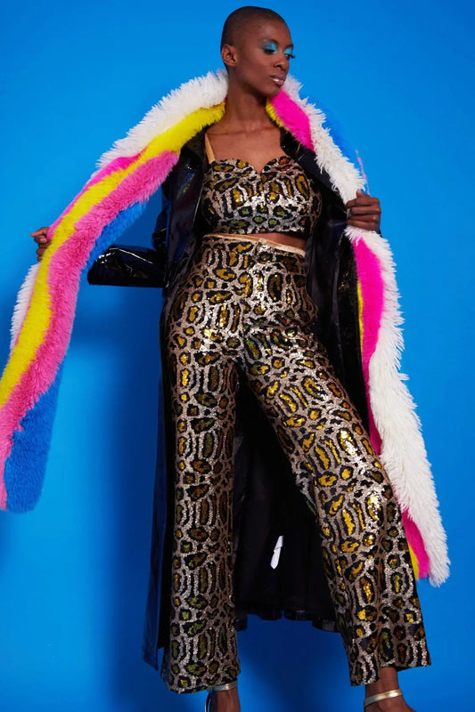 A person poses in a sequin leopard-print crop top and matching trousers, wearing a long black coat with a dove-grey faux fur snood, standing against a bright blue background.