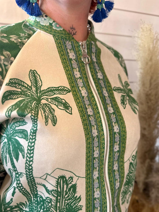 Person wearing a cream and green zip-up jacket with palm tree patterns and floral trim; blue tasseled earrings are partially visible.
