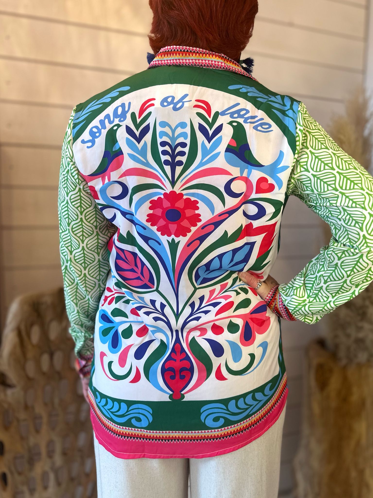Person with short red hair wearing a funky statement shirt with floral patterns and song of faith on the back, standing indoors facing away from the camera—perfect for festival season.