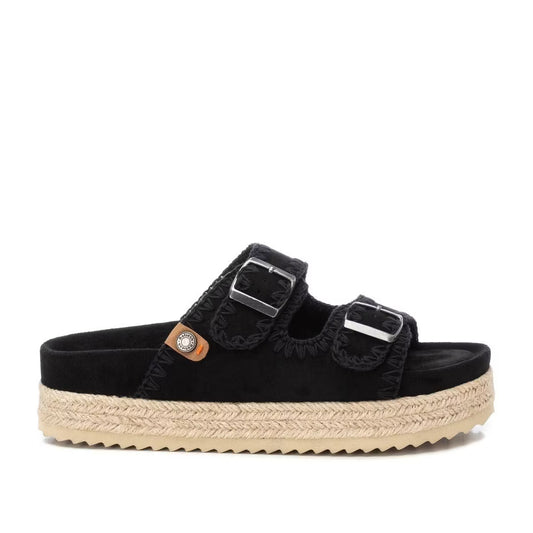 A black vegan suede sandal featuring two adjustable buckles, a woven jute platform sole, and a small round decorative detail on the side.