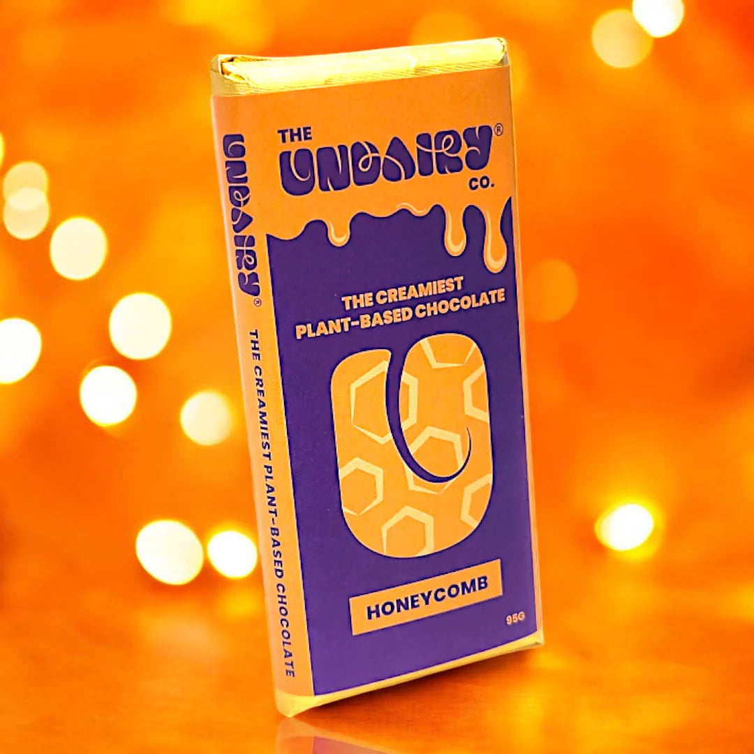 A 95g bar of The UnDairy Co. Vegan Non-Dairy chocolate in honeycomb flavour, with a blue and yellow wrapper, displayed against an orange background with blurred lights—an ethically conscious treat.