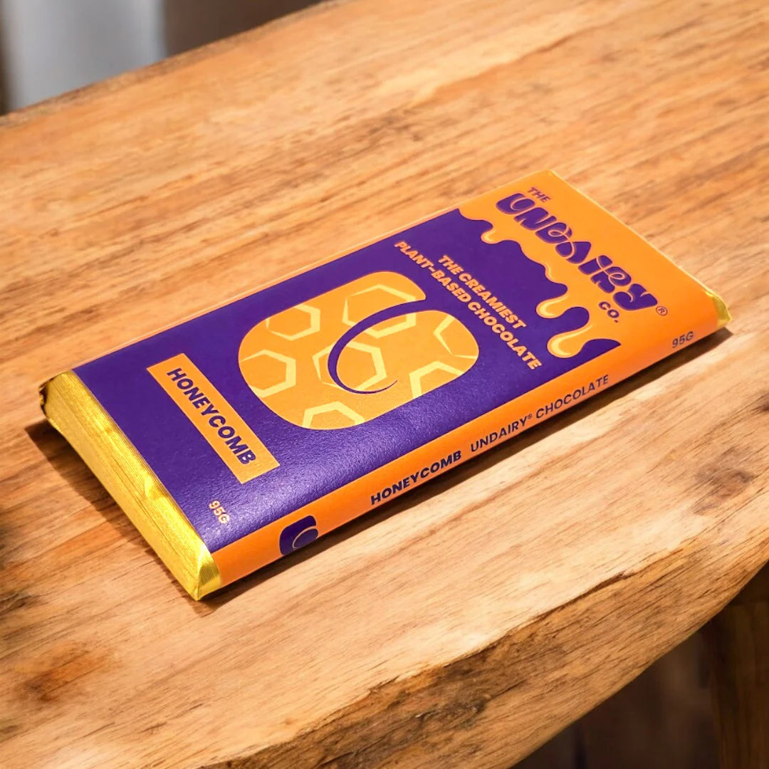 A chocolate bar labelled Honeycomb in purple and yellow packaging rests on a wooden surface. This ethically conscious treat is plant-based, vegan, and non-dairy chocolate, weighing 85 grams.