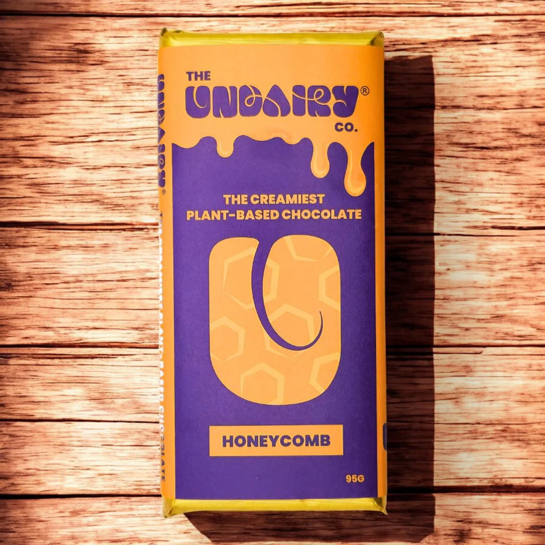 A 95g vegan non-dairy chocolate bar labelled The UnDairy Co. The Creamiest Plant-Based Chocolate, Honeycomb, rests on a wooden surface. The purple and yellow packaging features honeycomb patterns—an ethically conscious treat.