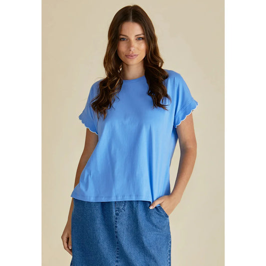 A woman stands against a beige background wearing the SLONE SCALLOP BLUE TEE and a blue denim skirt, embracing ethical fashion with one hand in her pocket.