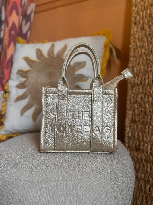 A small metallic tote bag labelled THE TOTE BAG rests on a textured grey seat with a sun-patterned cushion in the background.
