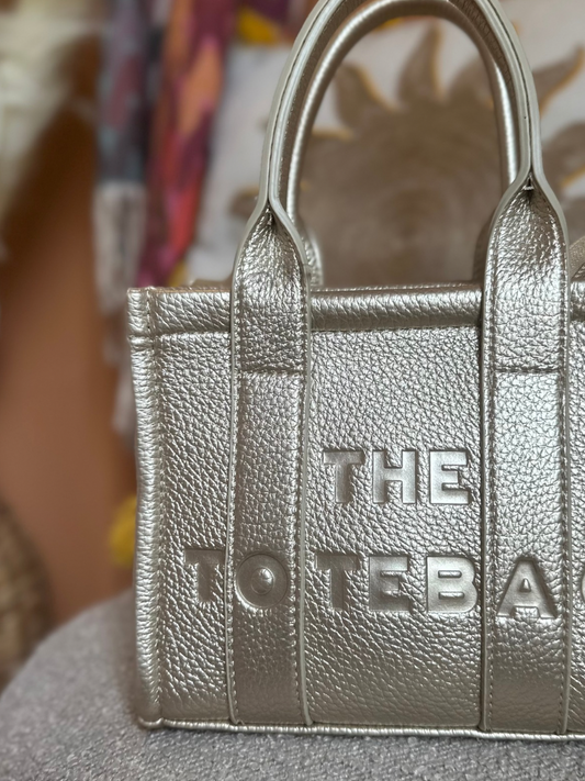 A metallic silver textured handbag with embossed lettering THE TOTE BAG partially visible on the front.