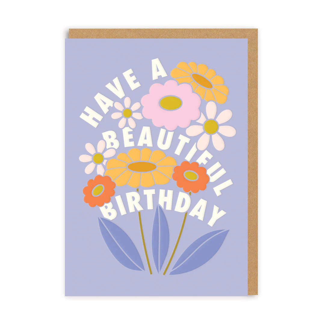 A birthday flower card featuring pastel blooms on a purple background, with the message 'Have a Beautiful Birthday' in white, elegantly curved around the cheerful flowers.