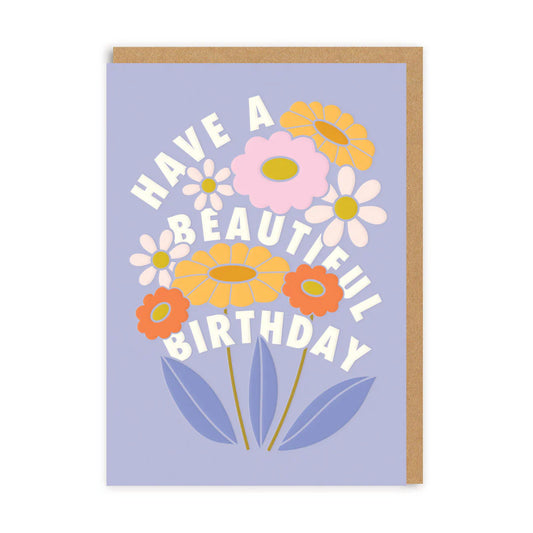 A birthday flower card featuring pastel blooms on a purple background, with the message 'Have a Beautiful Birthday' in white, elegantly curved around the cheerful flowers.
