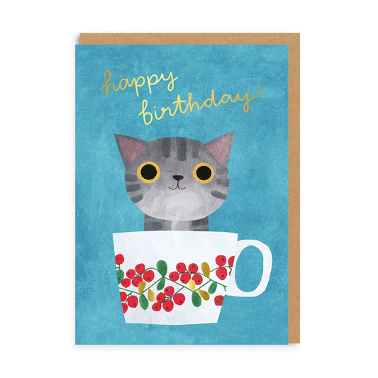 Whimsical birthday card featuring a grey cat with yellow eyes sitting in a floral teacup on a blue background. “Happy birthday!” is written at the top in cheerful yellow handwriting.
