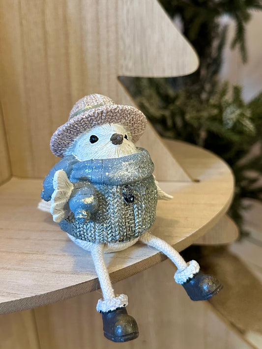 A charming ornament, this small decorative bird figurine wearing a blue jumper, boots, and a pink hat rests on a wooden shelf with a blurred tree visible in the background.