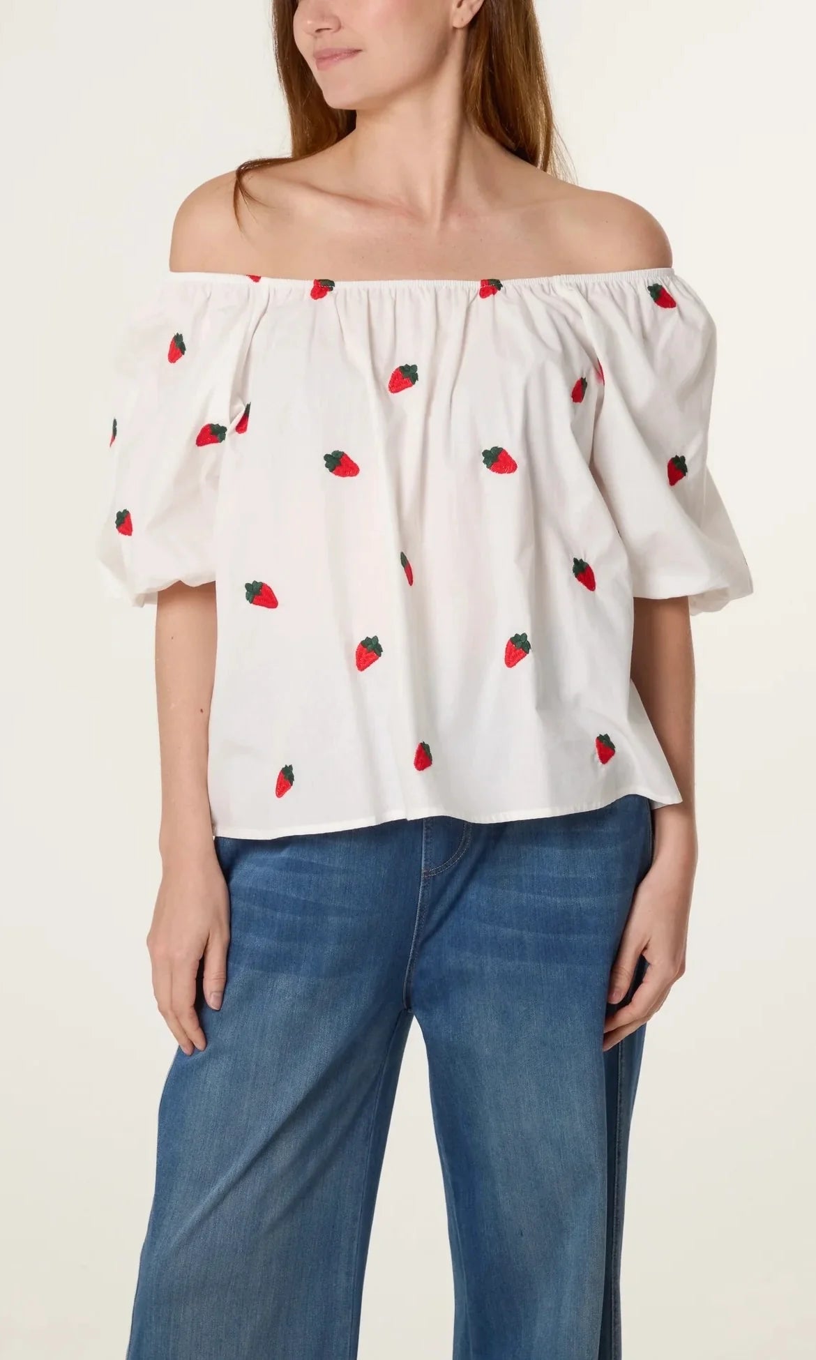 Woman in a Strawberry Ivory Puff Sleeve Top with off-the-shoulder puffy sleeves and a delightful strawberry print, stylishly teamed with blue jeans.