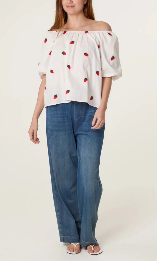 A woman wears a Strawberry Ivory Puff Sleeve Top featuring red strawberry prints, blue wide-leg jeans, and white sandals, standing against a plain backdrop.