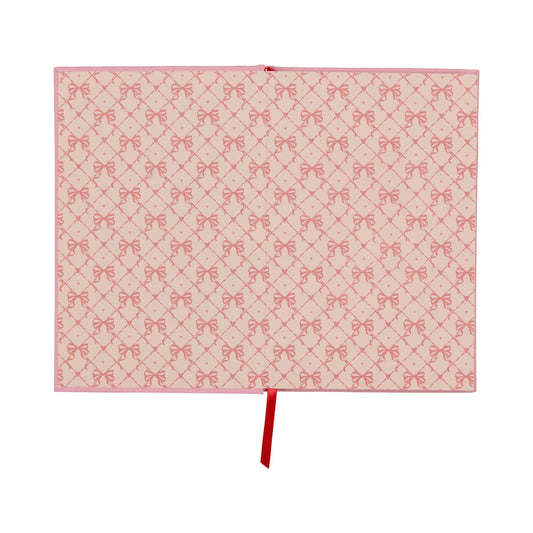 An open lay-flat journal with pink covers and pages displaying a repeating pattern of red bows, along with a red ribbon bookmark positioned in the centre.