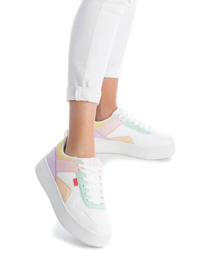 Person wearing low-top pastel trainers featuring vegan leather, with white trainers accented in pastel pink, green, and yellow, effortlessly paired with white rolled-up trousers.